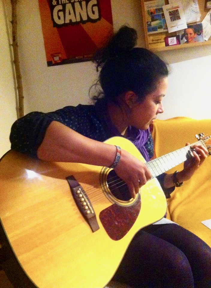 Junge Frau sitzt auf einem gelben Sofa und spielt an der Akustikgitarre